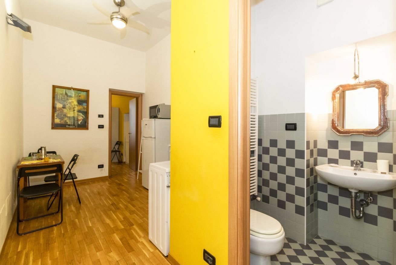 Una habitación con suelo de madera, pared amarilla, radiador blanco, inodoro blanco, lavabo con espejo y una pequeña mesa con sillas.
