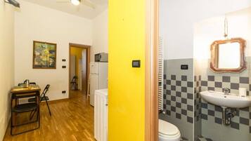 Una habitación con suelo de madera, pared amarilla, radiador blanco, inodoro blanco, lavabo con espejo y una pequeña mesa con sillas.