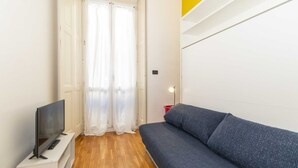 A room with a television on a stand, a sofa, a door with curtains, and a light fixture. - GuestHost - Porta Nuova Mini Flat 2 New! (Torino)