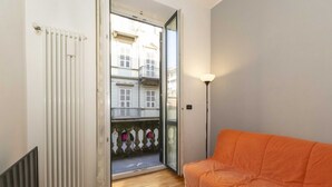 Una habitación con suelo de madera, un radiador blanco, un sofá naranja, un vidrio que da a un balcón y una lámpara.
