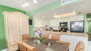 A kitchen with white cabinets, a black refrigerator, and a dining area with wicker chairs and a glass table. - Sand Dollar 1-504 - 5th Floor Ocean Front Condo in St. Augustine! (St. Augustine)