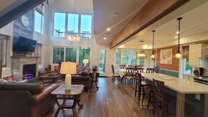 A spacious living room with a fireplace, a dining area with a long table and chairs, and a kitchen with a large island and pendant lights. - CAN-Luxury cottage by the water (Severn Bridge)