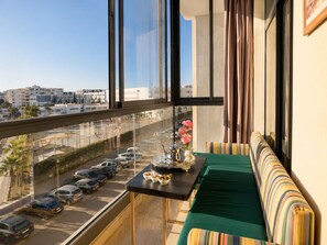 A balcony with a view of a parking lot and buildings, a sofa with striped cushions, and a small table with a vase of flowers. - Loli Home Agadir - Bay Premium Pool (Agadir)