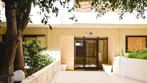 A building entrance with a glass door, a tree, and a white wall with a number sign.