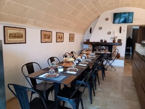 A dining area with a long table set for a meal, surrounded by chairs. There are framed pictures on the wall, a television mounted above the fireplace, and a kitchen area visible in the background.