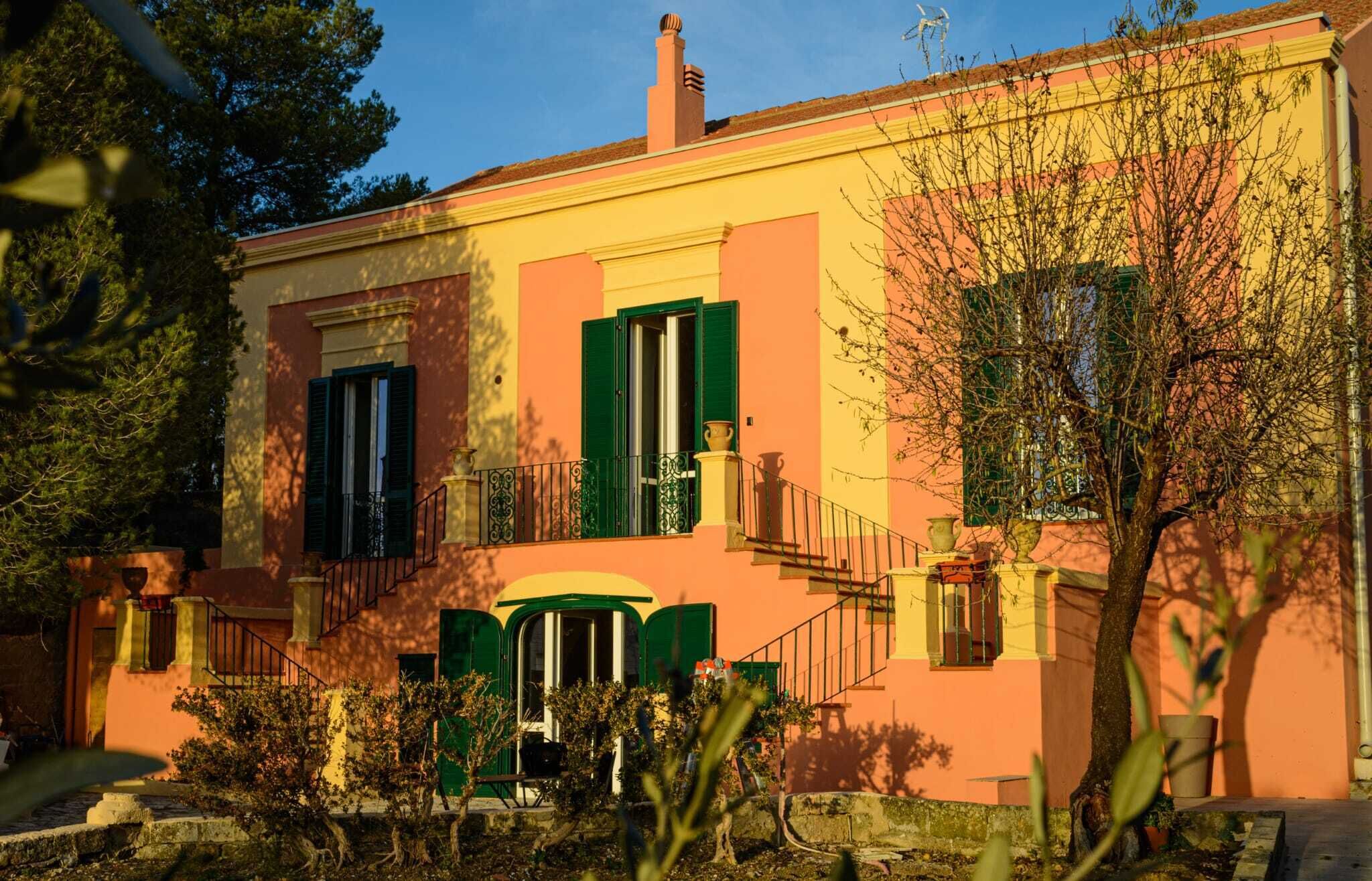 Un edifici de dues plantes amb façana groga, persianes verdes i un petit balcó.