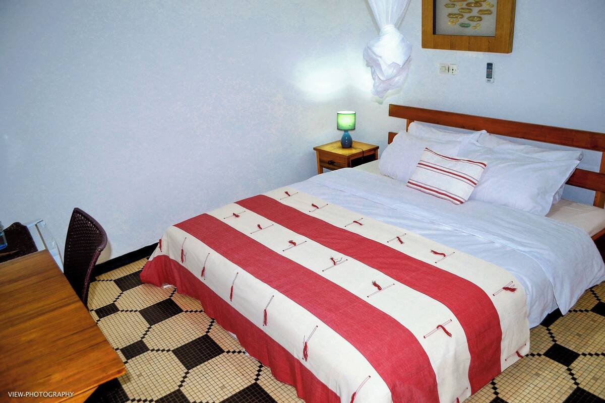A neatly made bed with a checkered floor, a bedside table with a lamp, and a wall-mounted power outlet.