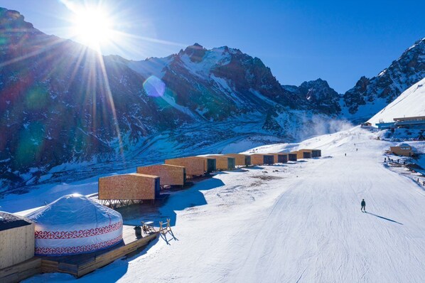 A snow-covered mountain with a clear sky and a sun shining. There are wooden cabins and a covered vehicle on the snow.