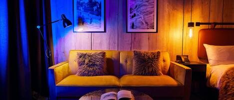 Quarto com sofá amarelo, duas imagens emolduradas na parede, mesa de cabeceira com abajur e mesa redonda com um livro aberto.