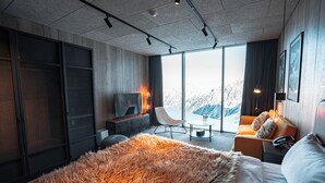 Ett modernt hotellrum med en stor säng, en sittgrupp med en fåtölj och ett bord, en TV och utsikt över bergen genom stora fönster.