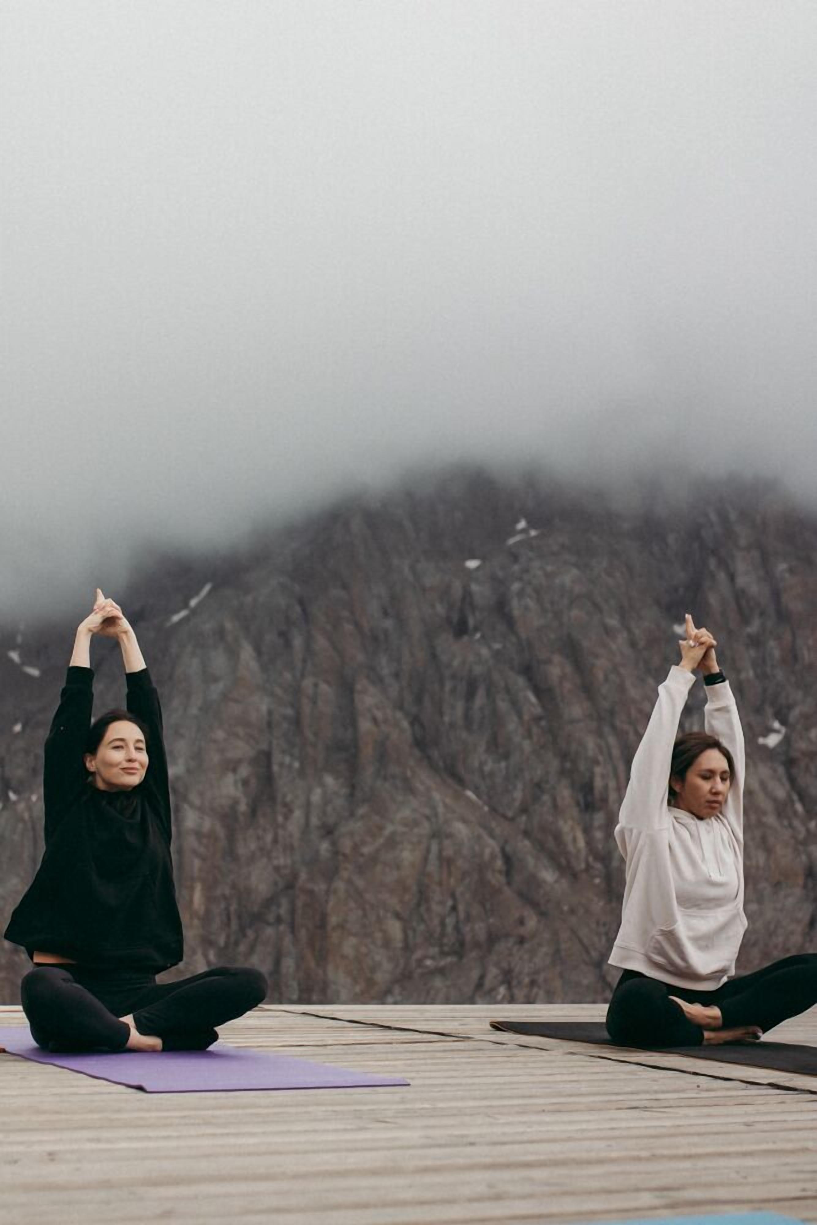 Zwei Personen praktizieren Yoga auf einer Holterrasse vor einer felsigen Bergkulisse.