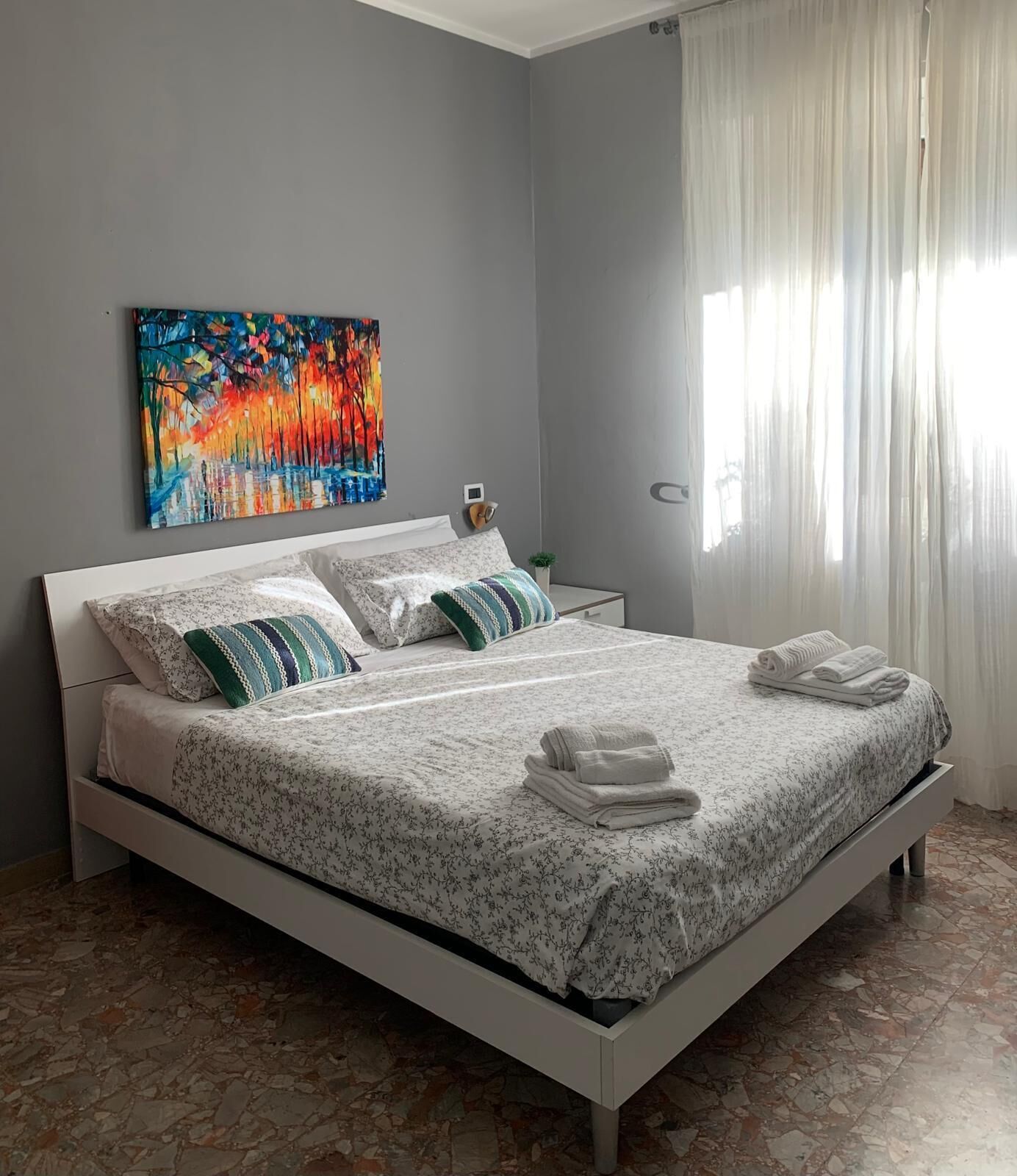 A bedroom with a bed, pillows, a painting, a nightstand, and a window with curtains.