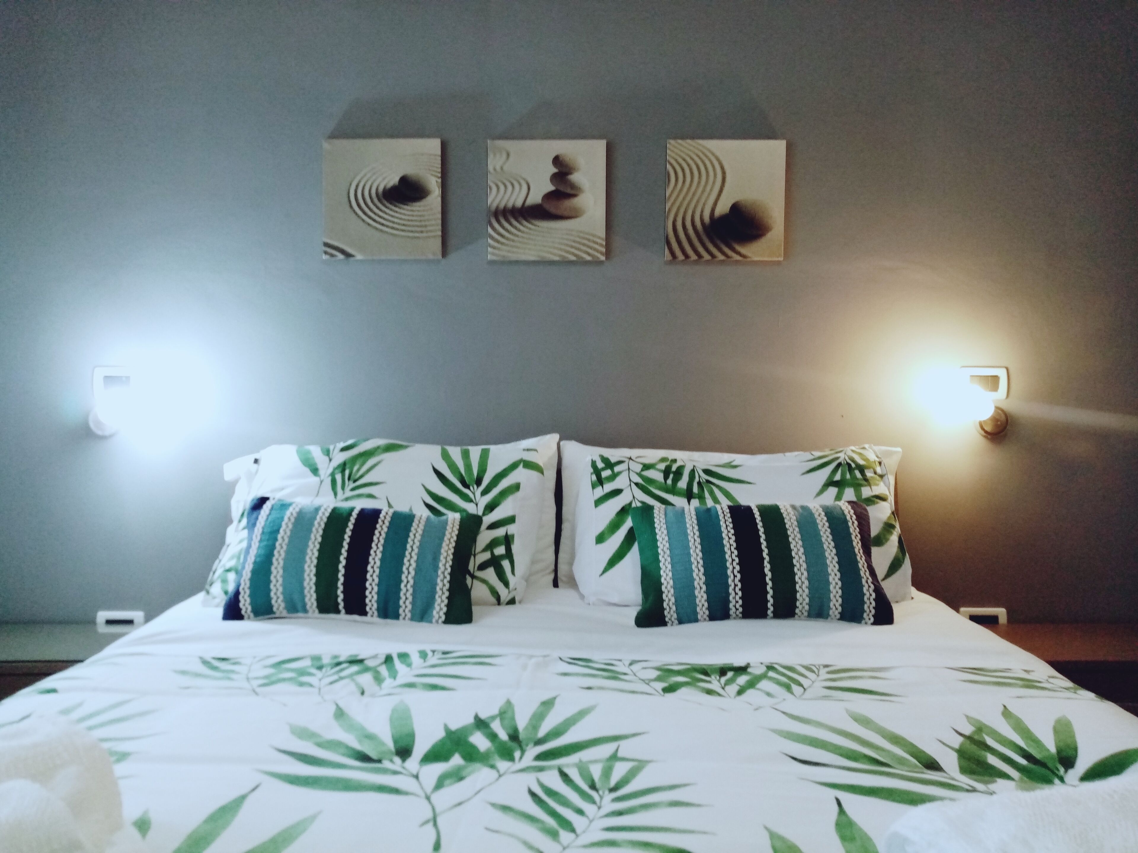 Un letto rifatto con cura, con una coperta a motivi fogliacei, due cuscini a righe e tre opere d'arte appese al muro sopra il letto.