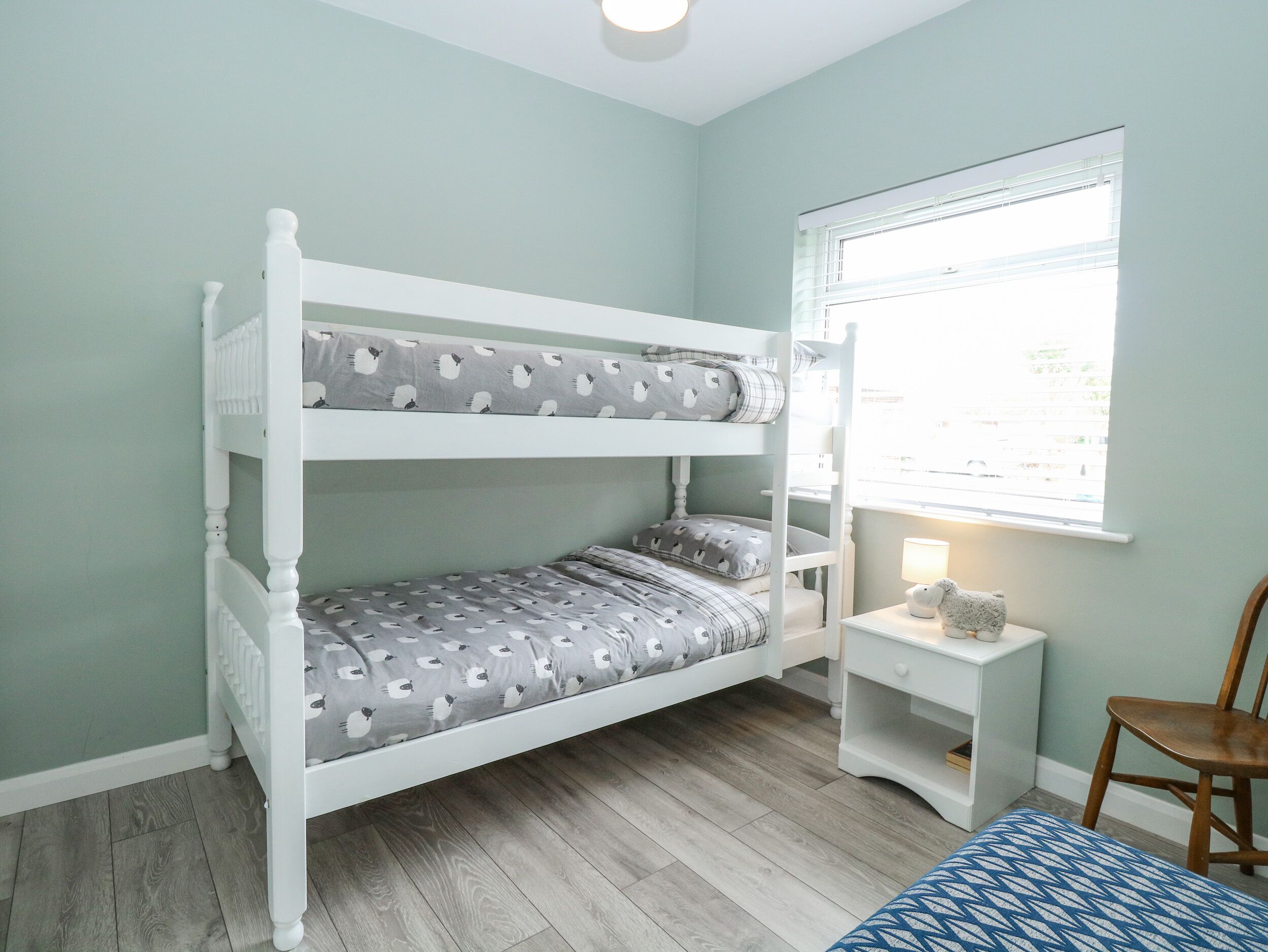 A bunk bed with grey and white patterned bedding, a small bedside table with a lamp, and a window with blinds.