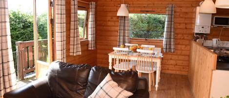 A cozy cabin interior with a leather sofa, plaid curtains, a dining table with chairs, and a kitchen area.