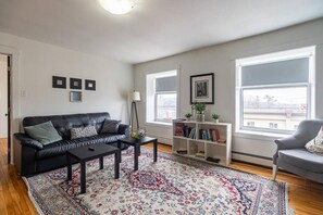 Una sala de estar con un sofá de cuero negro, una mesita de centro, un tapete estampado, una estantería y ventanas amplias.