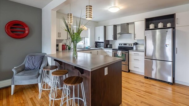 A modern kitchen with a central island, bar stools, stainless steel appliances, and a grey accent wall. - Large & Sunny Beautiful Home, AC (Bedford)