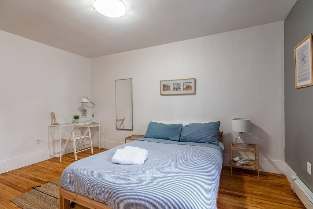 Un dormitorio con cama, escritorio, silla, mesita de noche y espejo en la pared.