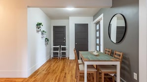 A dining area with a wooden table and chairs, a mirror on the wall, and a hallway with a door and plants. - Large & Sunny Beautiful Home, AC (Bedford)