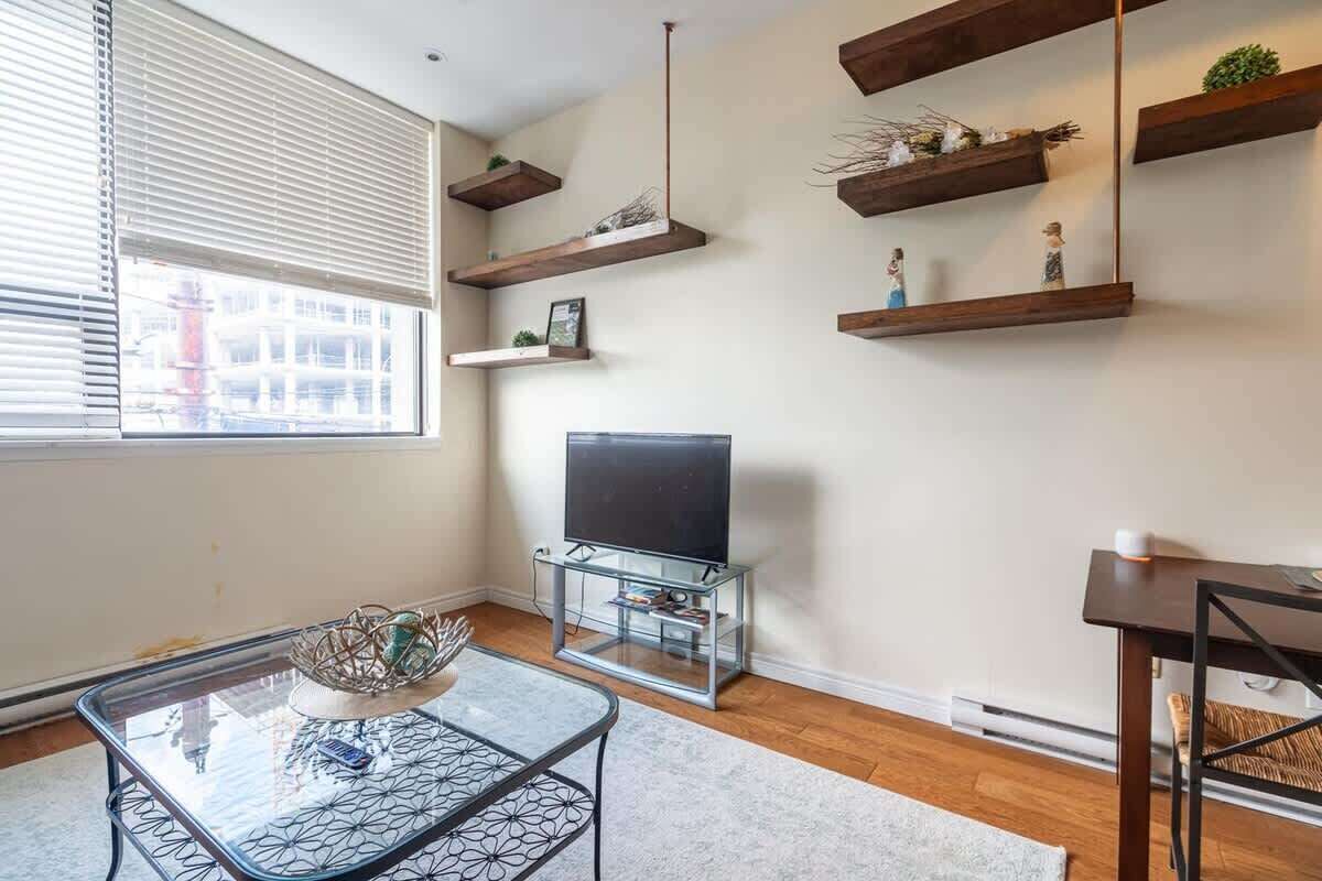 Un salon avec une table basse en verre, un téléviseur posé sur un meuble, des étagères en bois ornées d’objets décoratifs et une fenêtre avec des stores.
