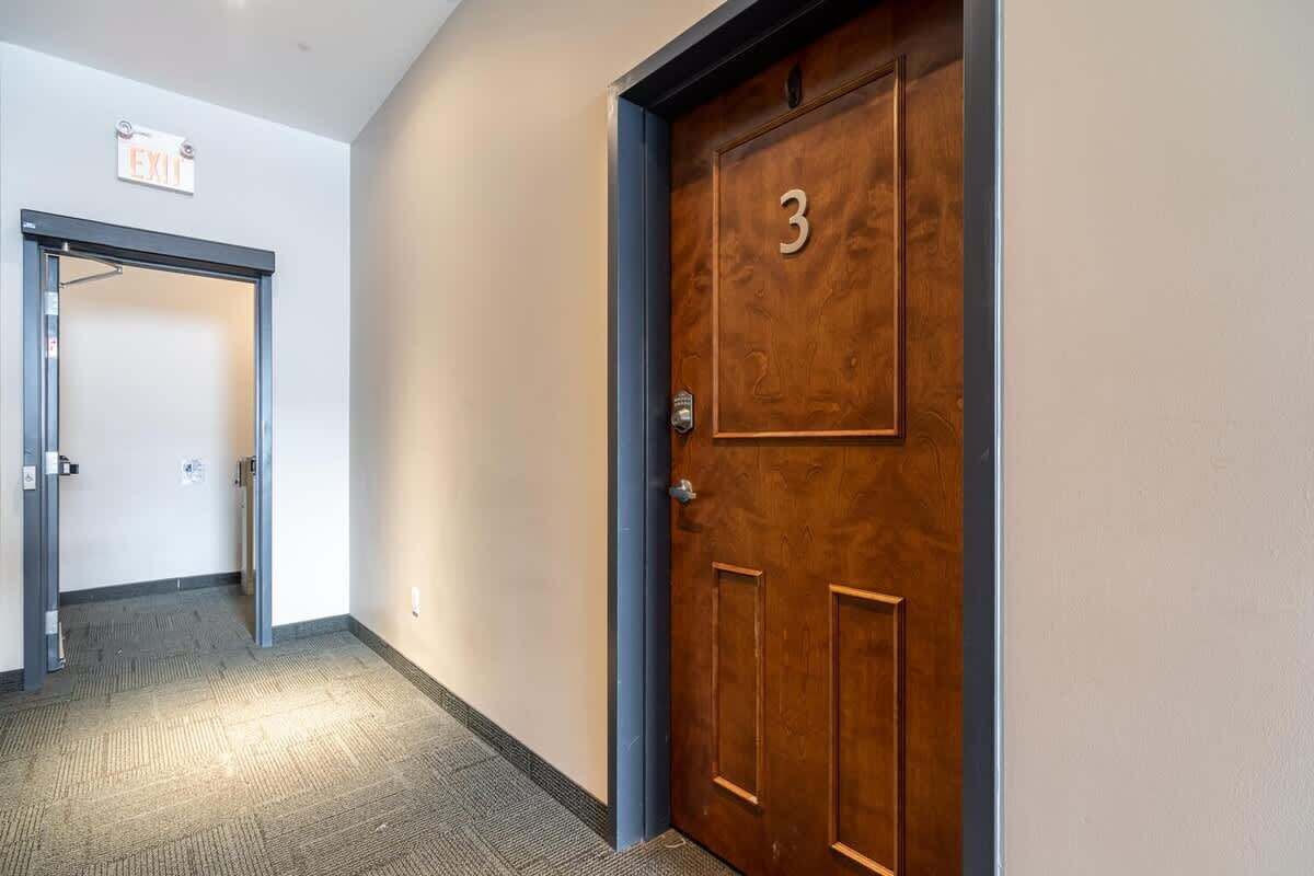 Un pasillo con una puerta de madera marcada con el número 3, un letrero de salida y suelo alfombrado.