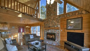 A log cabin interior with a stone fireplace, wooden furniture, and a large TV.