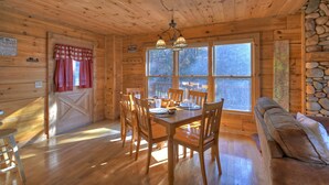 A wooden cabin interior with a dining table set for four, wooden walls, a stone wall, and a large window with a view of trees.