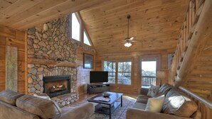 A log cabin interior with a stone fireplace, leather sofas, a television, and a balcony with a railing.