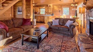 A log cabin interior with a wooden staircase, two leather sofas, a coffee table, a fireplace, and a kitchen area.