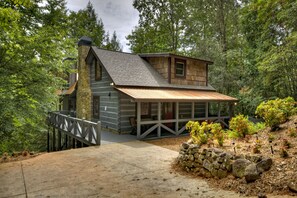A log cabin with a stone chimney, wooden deck, and a stone retaining wall. - Cozy Creek welcomes pets to its tranquil waters, offering fishing, swimming, a hot tub, outdoor fireplace, fire-pit, and WiFi (Epworth)