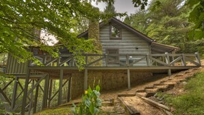 Une cabane rustique avec une cheminée en pierre, une terrasse en bois et un escalier menant à celle-ci.