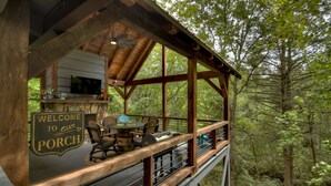 Une véranda en bois avec une pancarte de bienvenue, des sièges d’extérieur et un barbecue.