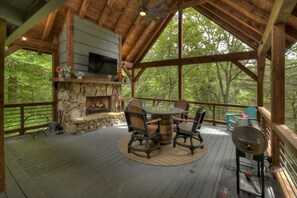 A covered outdoor deck with a stone fireplace, wooden furniture, and a grill. - Cozy Creek welcomes pets to its tranquil waters, offering fishing, swimming, a hot tub, outdoor fireplace, fire-pit, and WiFi (Epworth)
