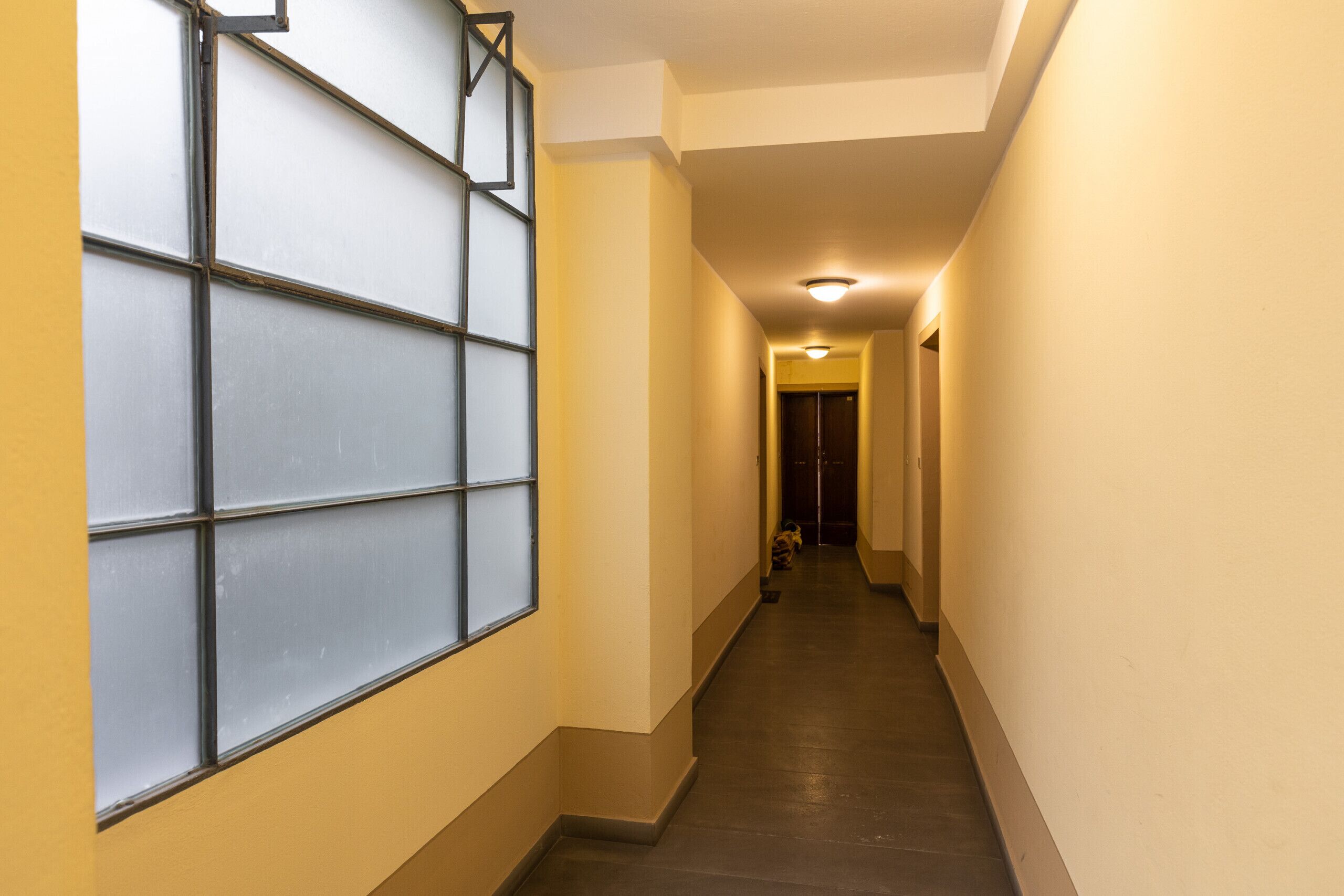 A narrow hallway with a tiled floor and a large window with a grid pattern on the left.
