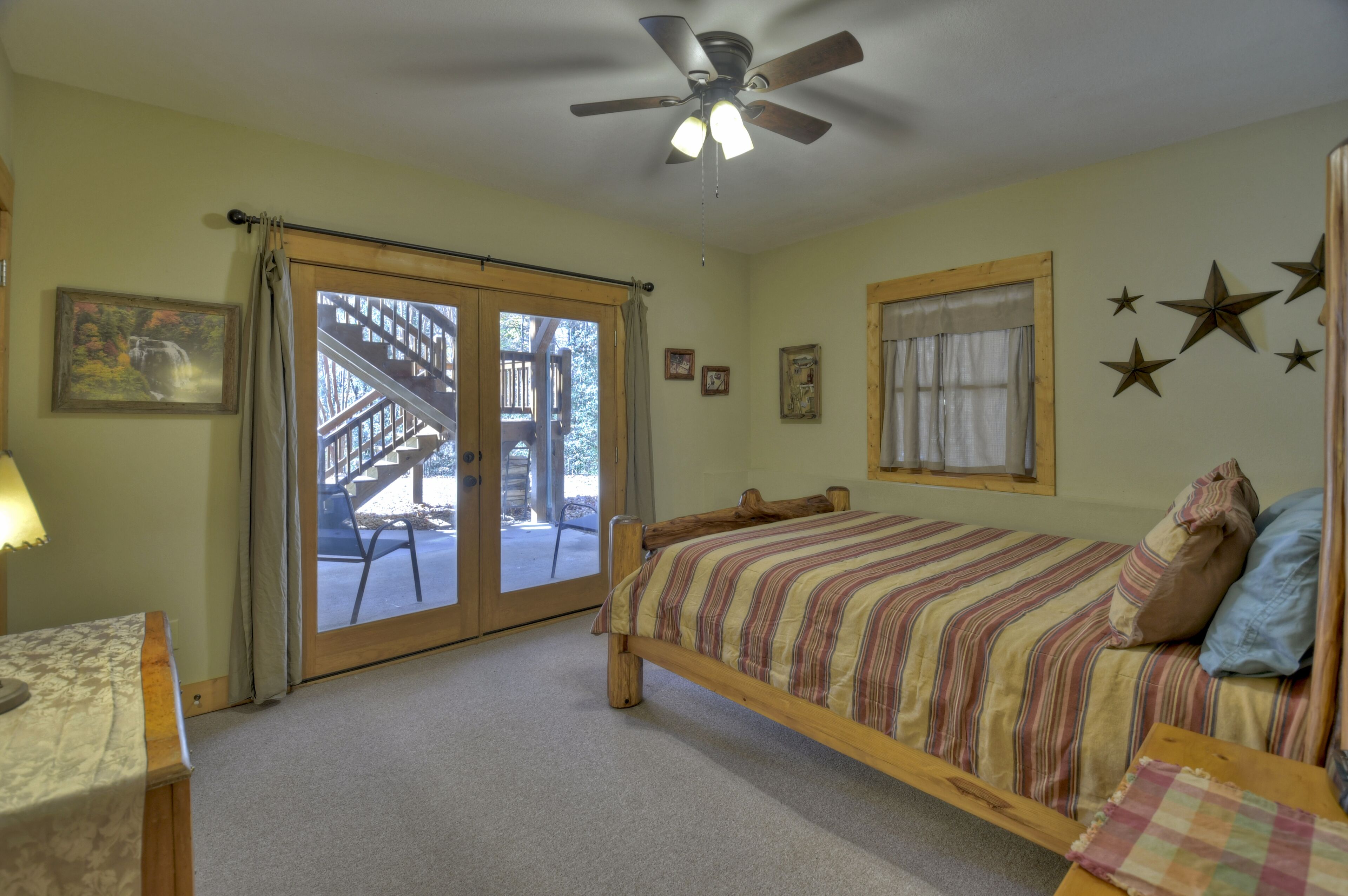 A bedroom with a bed, a ceiling fan, a window with curtains, and a view of an outdoor deck with a staircase.
