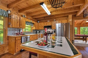 Une cuisine rustique aménagée dans une cabane en bois, avec un grand îlot central, des meubles en bois et une vue sur l’extérieur.