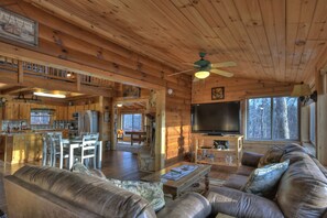 A cabin interior with a dining area, kitchen, living room with a sofa and TV, and a large window with a view of trees. - Discover Treetop Getaway -Pet-friendly retreat with panoramic mountain views, hot tub, firepit, fenced yard, WiFi, and a game-room (Ellijay)