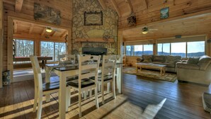 A spacious living area with a stone fireplace, wooden ceiling beams, and large windows offering a view of the outdoors.