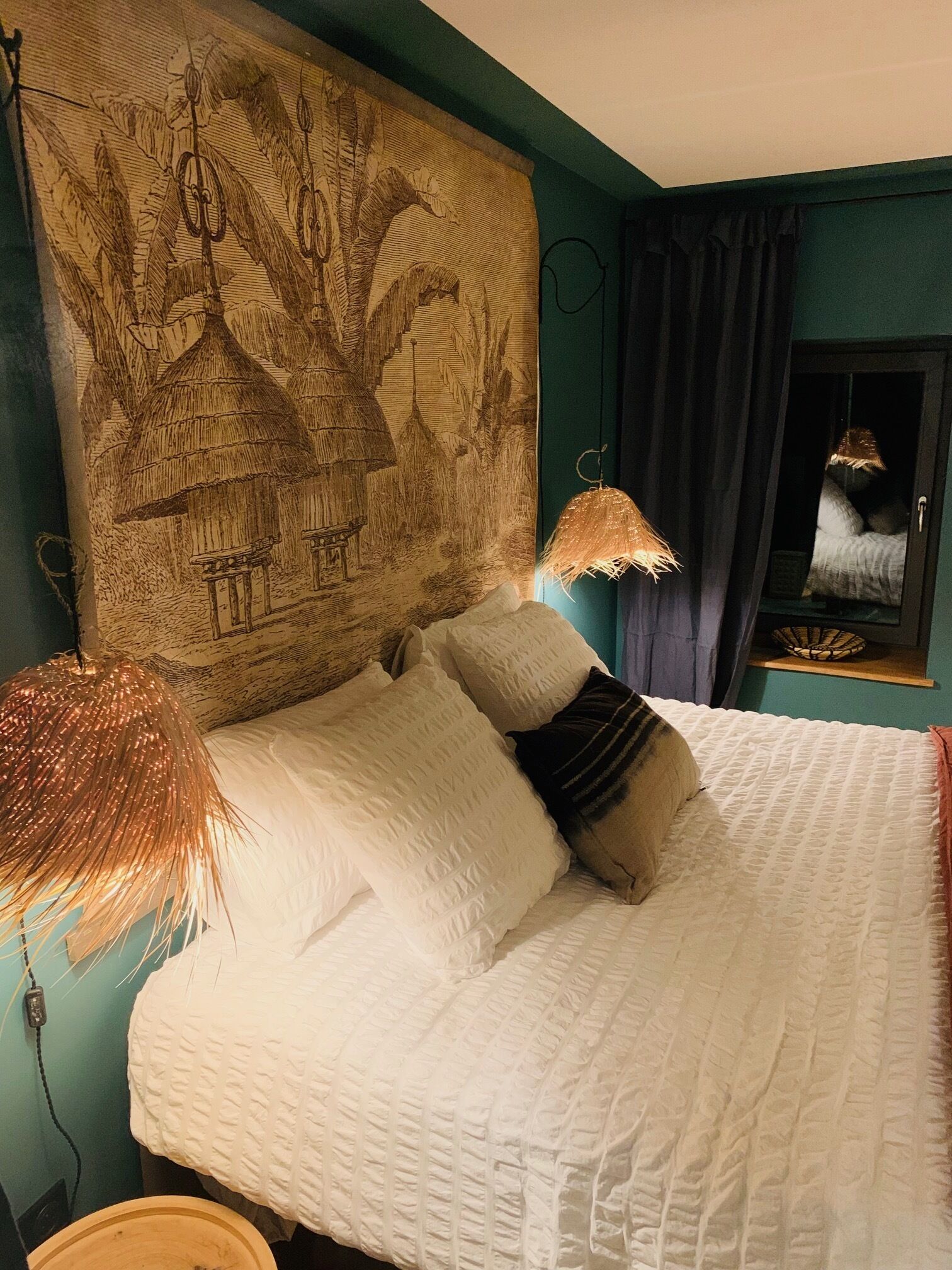 Een slaapkamer met een groot bed, een hoofdeinde met muurschildering, een hanglamp en een raam met gordijnen.