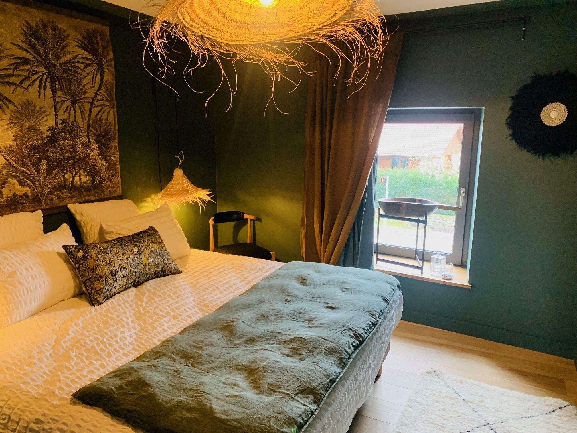 A bedroom with a bed, pillows, a hanging light fixture, a chair, and a window with a view.