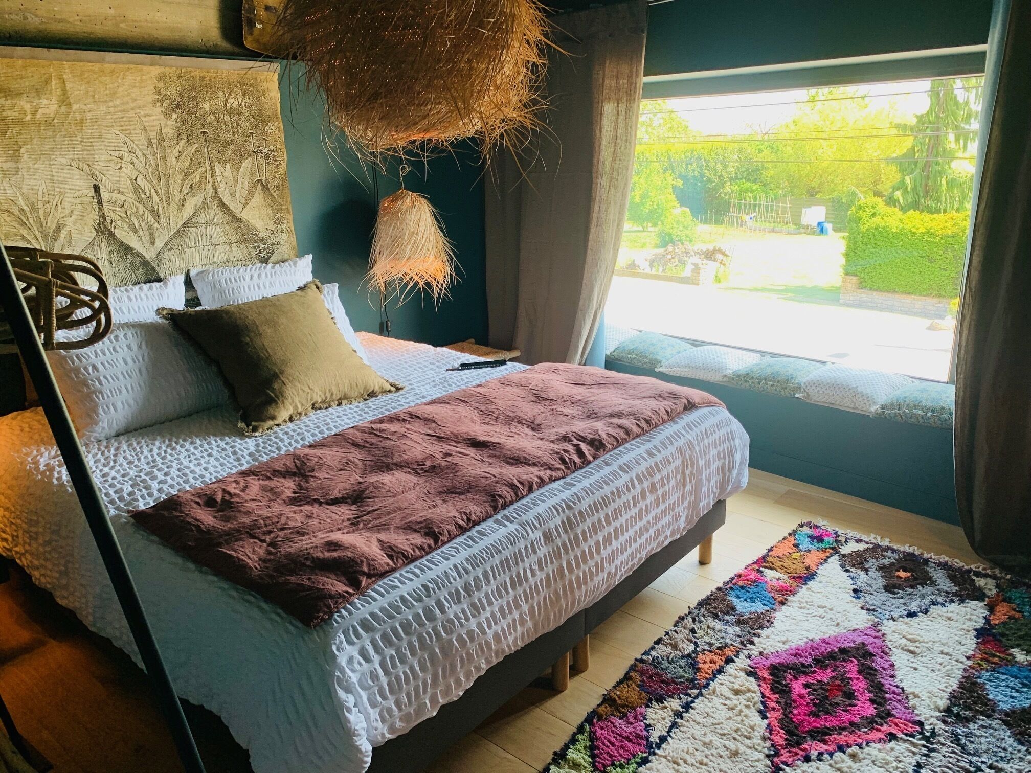 A bedroom with a bed, pillows, a hanging light, a window, and a view of greenery outside.
