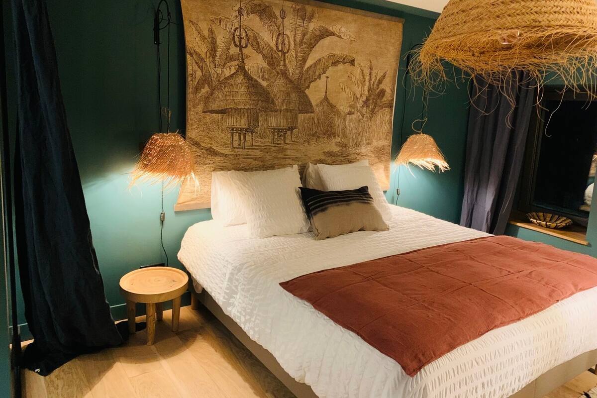 A bedroom with a bed, two bedside lamps, a small round table, and a large woven basket hanging from the ceiling.