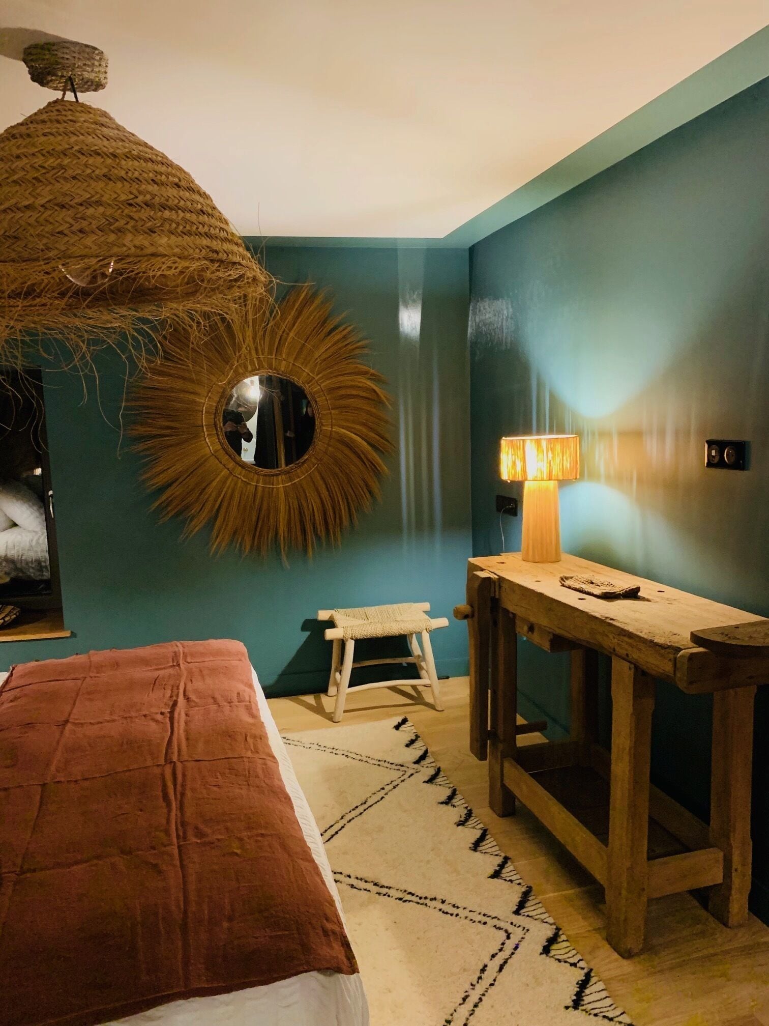 A room with teal walls, a round mirror, a hanging light fixture, a wooden bench, and a bed with a brown blanket.