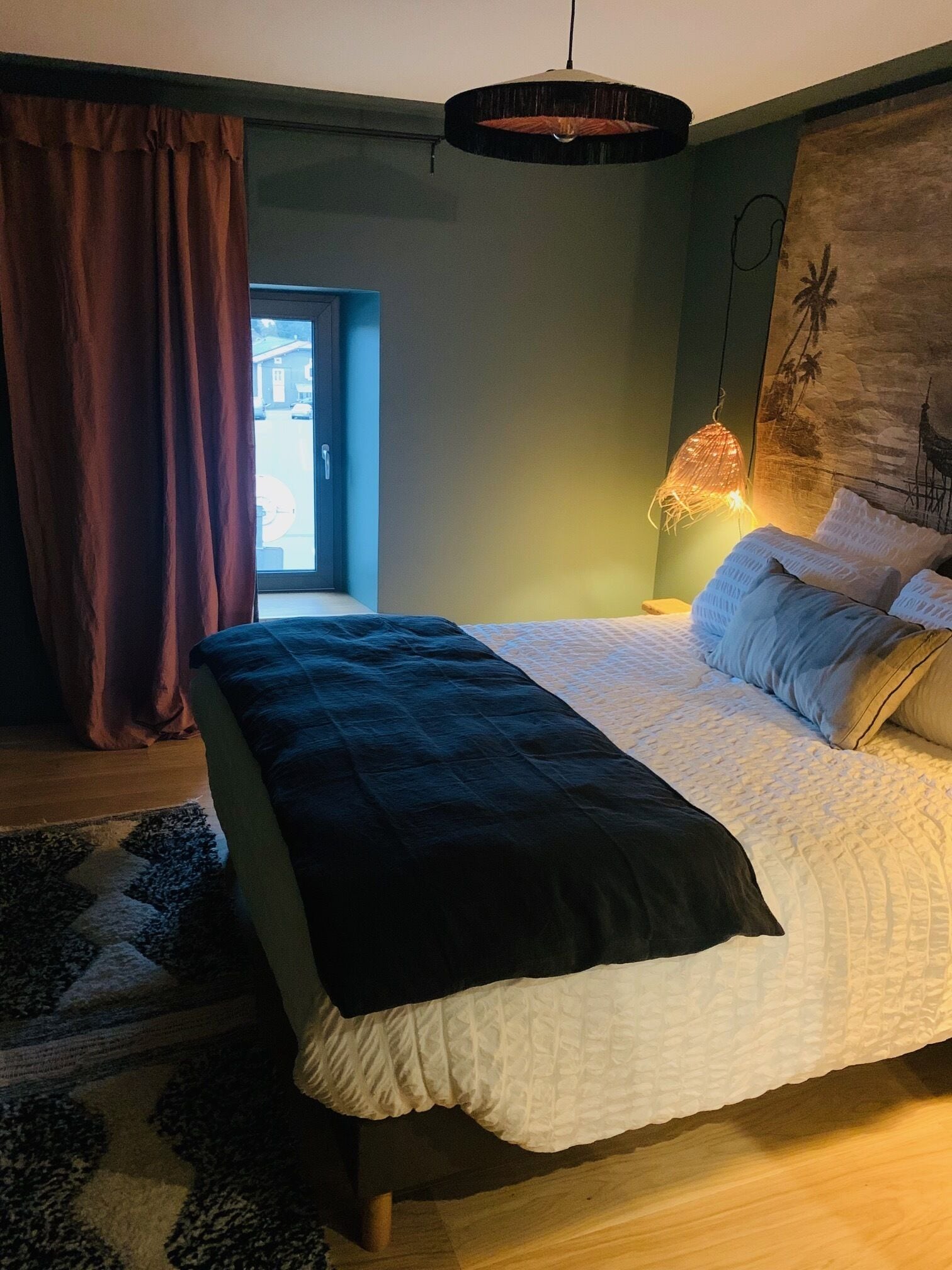 Une chambre à coucher avec un lit, un luminaire suspendu, un tableau au mur et une vue sur l’extérieur.