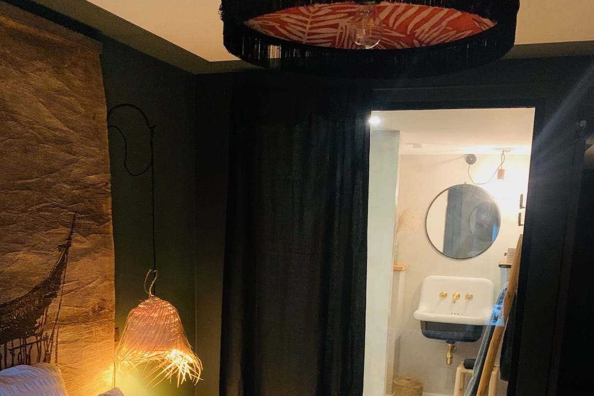 A bedroom with a bed, a hanging light fixture, and a view into a bathroom with a sink and mirror.