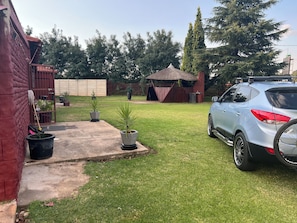 A backyard with a car parked on the right, a thatched-roof structure, and potted plants. - Fabulous Airbnb for Leisure and Business Comfort (Springs)