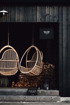 Des chaises en osier suspendues à un plafond en bois, une enseigne indiquant « HILLTOP HOUSE », et un panneau d’accueil qui dit : « ENTREZ, NOUS SOMMES OUVERTS ».