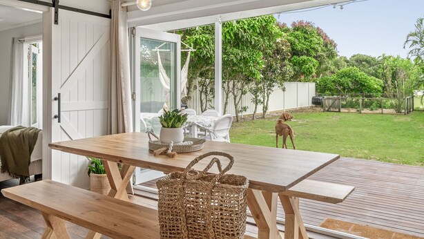 Een houten terras met een tafel en stoelen, een rieten mand en een hond in een achtertuin met een gazon en bomen.