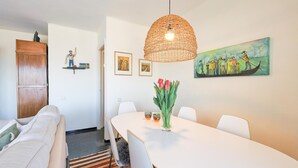 Een eetruimte met een witte tafel, stoelen, een geweven hanglamp, een vaas met rode tulpen en een schilderij van een bootscène.