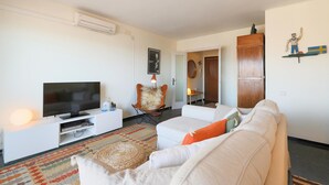 Een moderne woonkamer met een flatscreen televisie, een witte bank met oranje kussens, een kamerbreed tapijt en een houten kast.